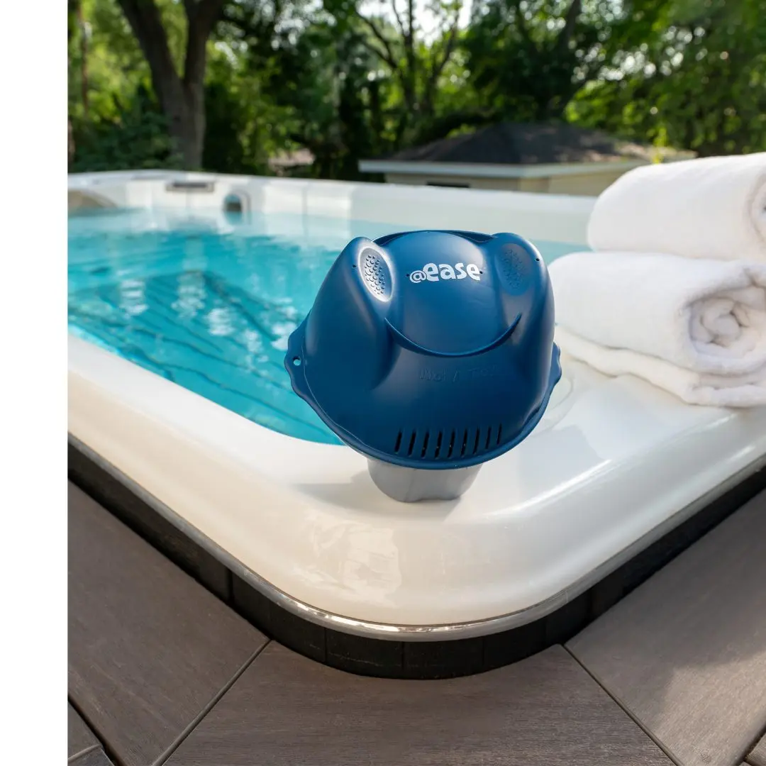 A FROG @ease Sanitizing System for Swim Spas sits on the edge of an in-ground swim spa alongside some rolled up white towels