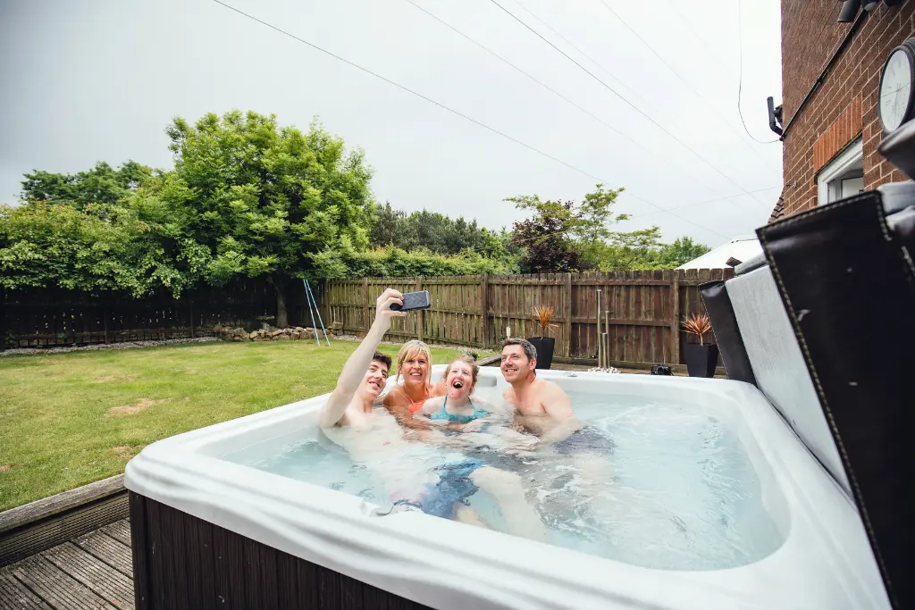 Four friends taking a selfie in an outdoor hot tub on a backyard deck.
