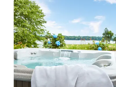 An outdoor hot tub is photographed with a lake and greenery in the background. The water is clear and there is a white towel hanging off the front edge of the spa.