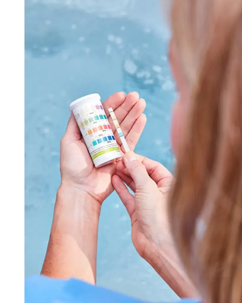Blonde woman holding up a colored FROG Test Strip and comparing it to the colors on the bottle